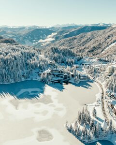 Luftaufnahme des Hotel Hochschober am zugefrorenen Turracher See mit verschneiten Hängen und Wald.