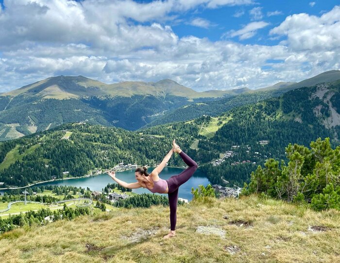 Eine Frau hält auf einer Bergwiese eine Yoga-Position mit Blick über die Turracher Höhe und den See.