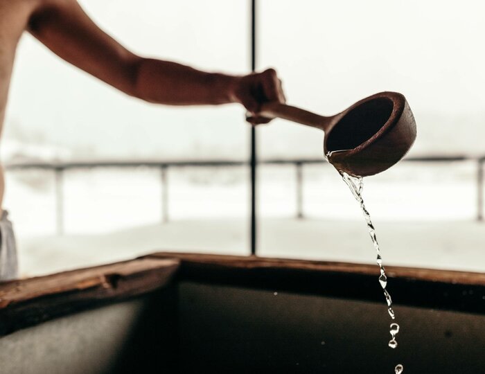 Ein Arm gießt Wasser aus einem Holzlöffel auf die heißen Saunasteine.