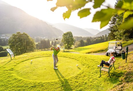 Ein Golfer schlägt auf einem sonnigen Abschlag ab, während eine Person daneben auf einer Bank sitzt.