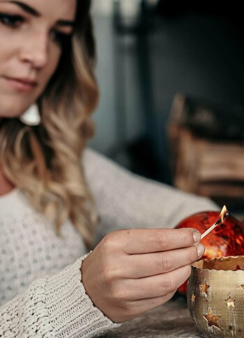 A woman lights a match at a table for a red Advent decoration in a gold-toned bowl.