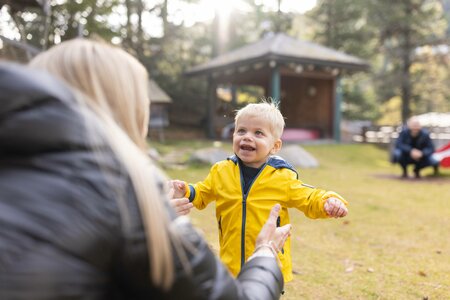 Ein Kleinkind im gelben Jacke lacht einer erwachsenen Person im Freien entgegen, im Hintergrund stehen Pavillons und weitere Gäste.