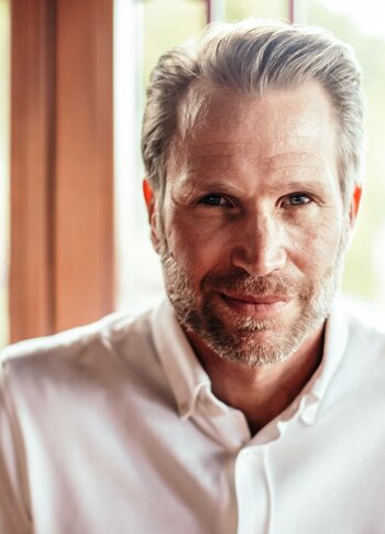 Ein Mann mit weißem Hemd blickt in die Kamera vor hellem Hintergrund.