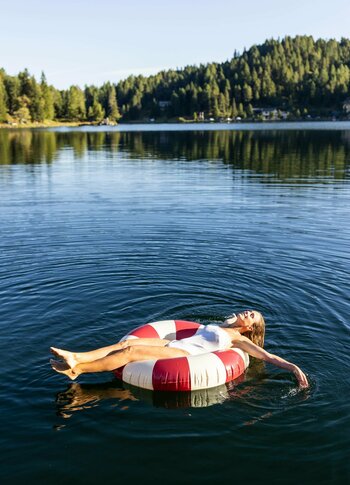 Eine Person liegt entspannt auf einem roten und weißen Schwimmreifen im ruhigen Wasser eines Sees. Umgeben von grünen Bäumen und sanften Hügeln, reflektiert das Wasser die klare blaue Himmel. Die Atmosphäre ist friedlich und einladend.