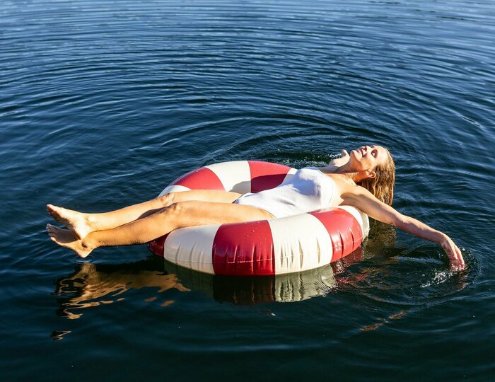 Eine Person liegt entspannt auf einem roten und weißen Schwimmreifen im ruhigen Wasser eines Sees. Umgeben von grünen Bäumen und sanften Hügeln, reflektiert das Wasser die klare blaue Himmel. Die Atmosphäre ist friedlich und einladend.