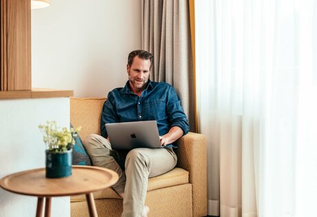 Ein Mann sitzt in einem Hotelzimmer auf dem Sessel und arbeitet am Laptop neben einem kleinen Holztisch.
