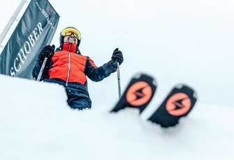 A skier in a red jacket stands at the start and holds ski poles in snowy terrain.