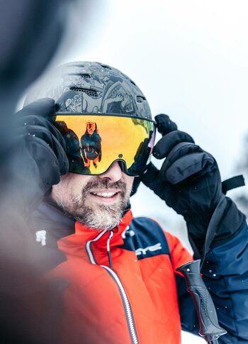 A skier adjusts his goggles while wearing a red jacket and gloves in the snow.