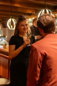 Eine Frau hält an der Bar ein Glas Weißwein und spricht mit einem Mann im Vordergrund.