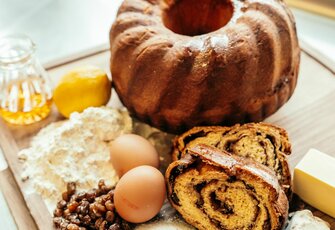 Auf einem Holzbrett stehen Gugelhupf, aufgeschnittene Roulade, Eier, Mehl, Nüsse, Butter und eine kleine Honigkanne.