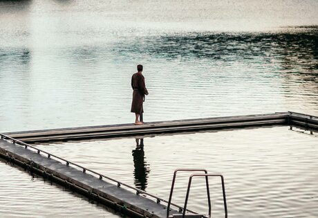 Eine Person steht auf dem Steg am See-Bad und blickt über das ruhige Wasser.