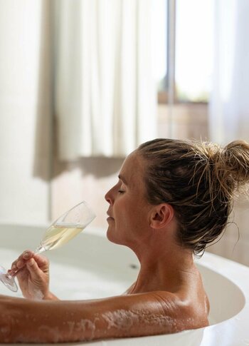 Frau liegt in einer Badewanne mit Schaum und trinkt Champagner.