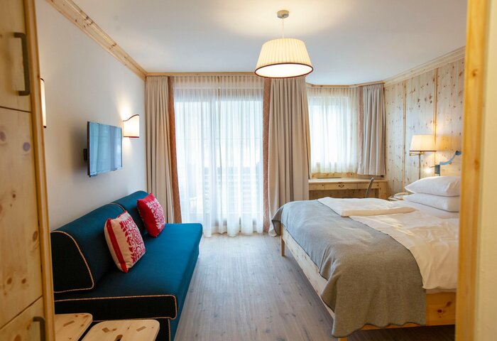 Ein Superior-Zirbenzimmer mit Bett, blauem Sofa, Holzmöbeln und großen Fenstern mit hellem Vorhang.