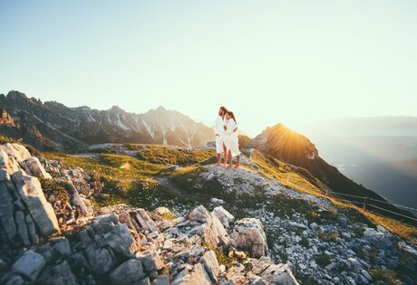 Zwei Personen stehen auf einem felsigen Berggrat in der Morgensonne vor einer Alpenkulisse.