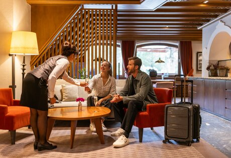 Eine Mitarbeiterin begrüßt zwei Gäste in der Hotelhalle; Koffer steht neben dem roten Sessel.
