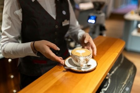Eine Servicekraft stellt eine Tasse Cappuccino auf der Holztheke an der Hotelbar ab.