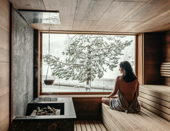 Eine Frau sitzt in der Holzsauna und blickt durch das große Fenster auf verschneite Bäume draußen.