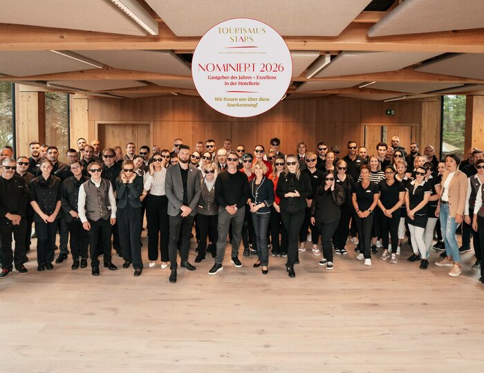 Ein großes Hotelteam posiert in einem hellen Raum vor einer Holzwand für ein Gruppenfoto.