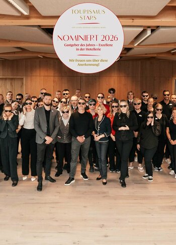 Hotelteam beim Gruppenfoto im Hotel Hochschober Ein großes Hotelteam posiert in einem hellen Raum vor einer Holzwand für ein Gruppenfoto.