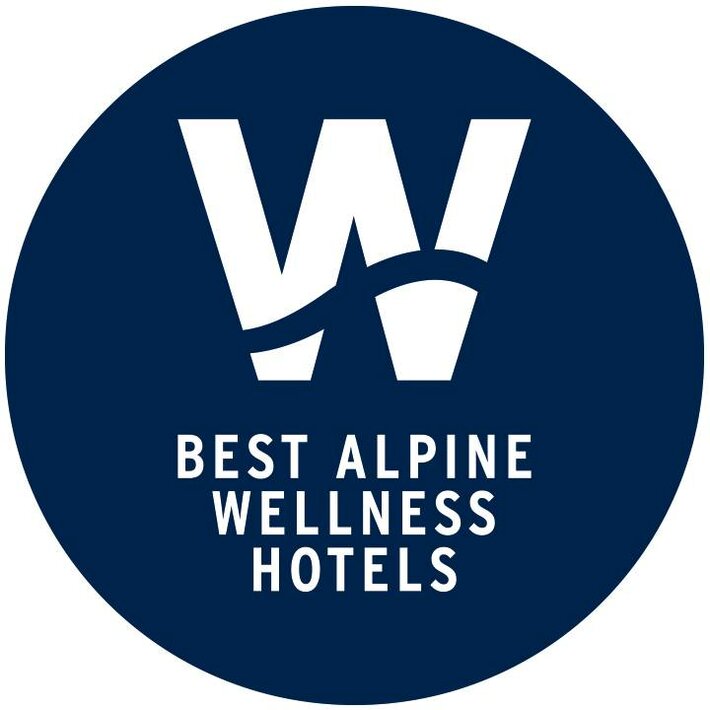 Ein rundes blaues Logo mit weißem W und der Aufschrift Best Alpine Wellness Hotels.