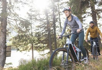 Zwei Mountainbiker fahren auf einem Waldweg am Seeufer entlang, die Sonne scheint durch die Bäume.
