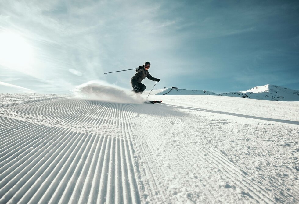 A skier carves across a freshly groomed slope with a snowy mountain backdrop.