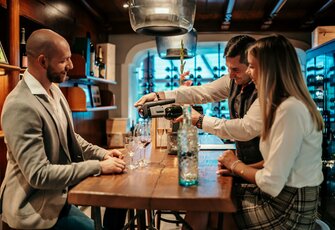 Drei Gäste an der Hotelbar, während ein Mann Getränke zubereitet und Weinflaschen im Hintergrund stehen.