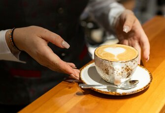 Eine Hand serviert eine Cappuccino-Tasse mit Latte-Art auf einem Unterteller am Holztisch.