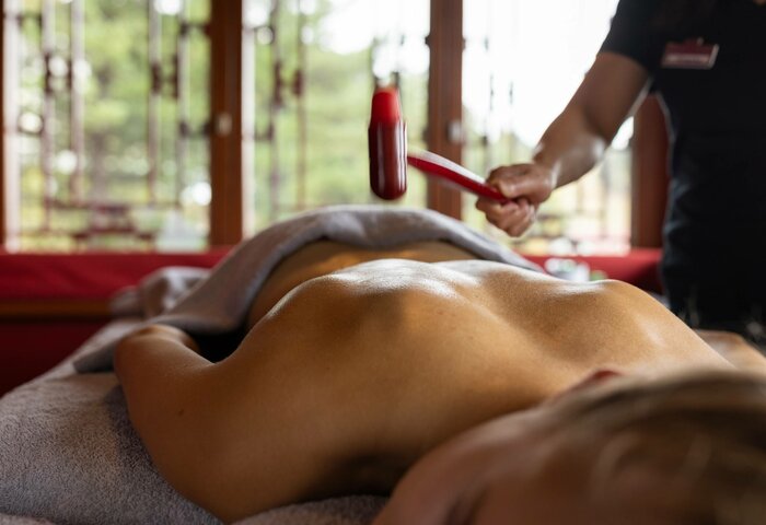 Eine Person erhält eine Massage auf einer Liege, während im Hintergrund eine Behandlerin mit einem roten Massagegerät arbeitet.