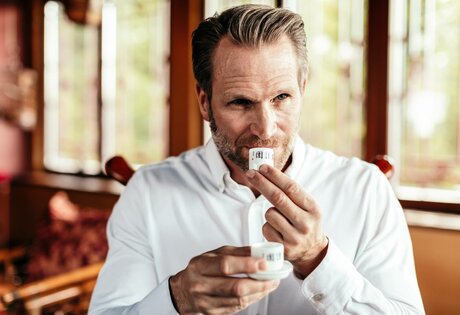 Ein Mann in weißem Hemd riecht an einer Teetasse und hält eine weitere Tasse in einem warm eingerichteten Raum.