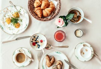 Ein Frühstückstisch mit Croissants, Eiern, Joghurt, Kaffee und Saft ist von oben arrangiert.