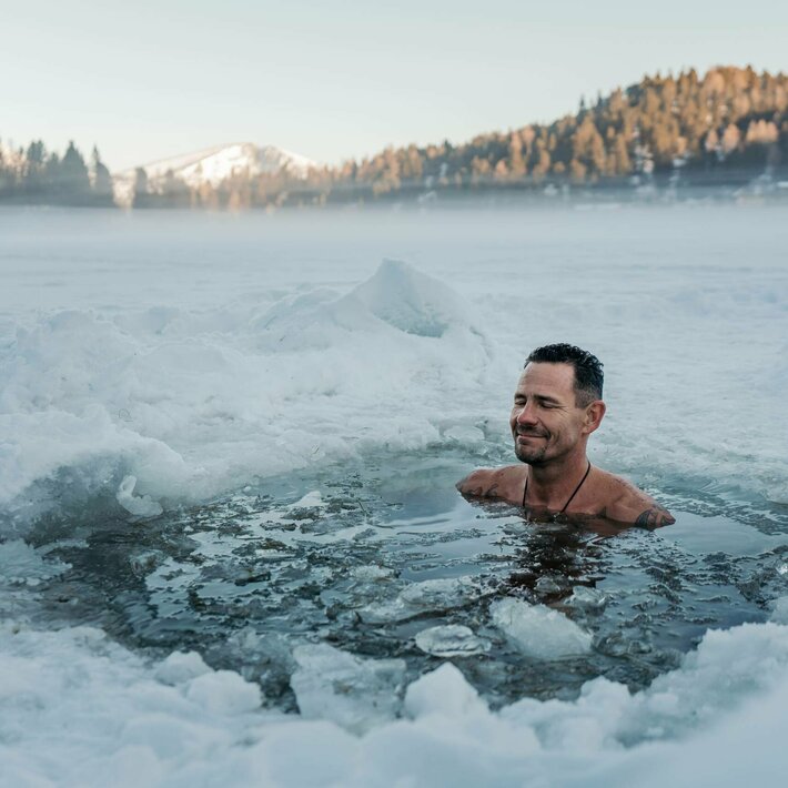 A man stands chest-deep in the icy water of the heated lake pool, surrounded by snow and mist.