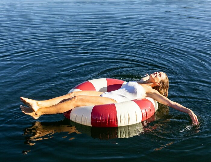 Urlaubsangebote für das ganze Jahr - Turracher Höhe Eine Person liegt entspannt auf einem roten und weißen Schwimmreifen im ruhigen Wasser eines Sees. Umgeben von grünen Bäumen und sanften Hügeln spiegelt sich die Natur im klaren Wasser wider. Die Atmosphäre ist friedlich und sonnig.