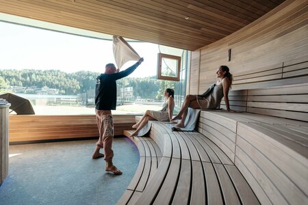 Neuheiten ab Sommer 2026 im Hotel Hochschober auf der Turracher Höhe in Kärnten. Drei Personen in einer modernen Sauna mit Holzverkleidung. Eine Person zieht einen Vorhang auf, während zwei andere entspannt auf den Bänken sitzen. Große Fenster bieten einen Blick auf eine bewaldete Landschaft. Tageslicht strömt in den Raum.