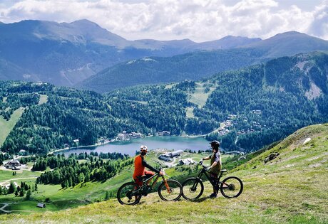 Zwei Mountainbiker stehen auf einer grünen Almwiese mit Blick auf den Turracher See und die Berge.