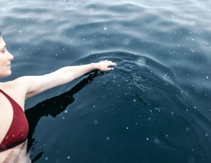 Wellnessbad im See beim Hotel Hochschober Eine Frau im roten Badeanzug schwimmt in dunklem Wasser und berührt die Oberfläche mit dem Arm.