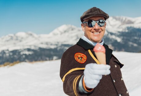 Ein Mann in Winterkleidung hält im Schnee eine Eiscreme vor verschneiten Bergen.
