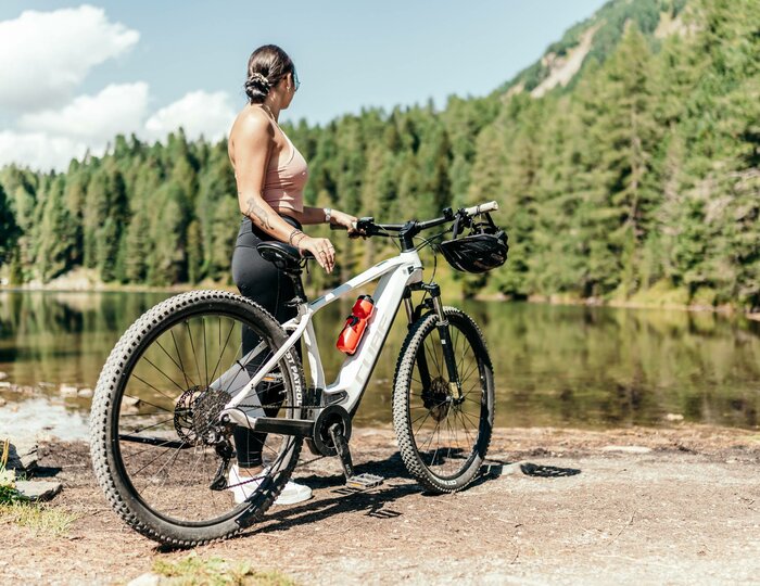 E-Bike-Ausflug am See auf der Turracher Höhe Eine Frau steht mit einem E-Bike am Ufer eines Bergsees vor Wald und Felsen.