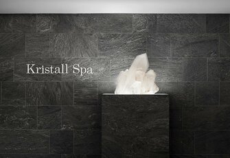 Ein weißer Kristall steht vor einer dunklen Steinwand mit der Beschriftung „Kristall Spa“.