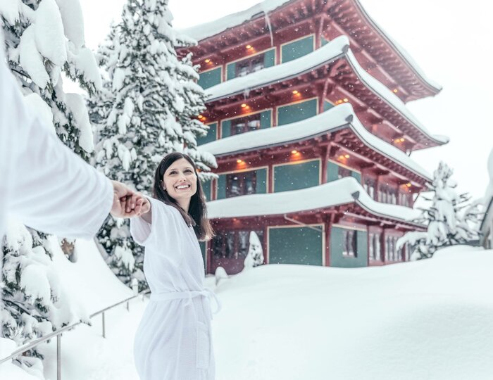A woman in a white bathrobe stands in a deep snowy outdoor area in front of the Chinaturm at Hotel Hochschober.