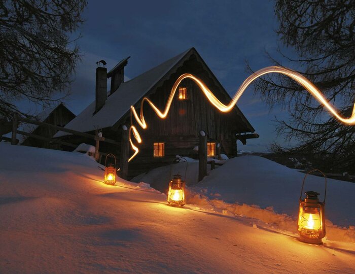 Bergweihnacht am verschneiten Berghaus Ein verschneites Berghaus bei Nacht, mit Laternen im Schnee und einer warmen Lichterlinie am Dach.