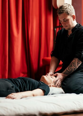 Ein Therapeut behandelt eine liegende Person mit Shiatsu auf einer hellen Liege vor roten Vorhängen.