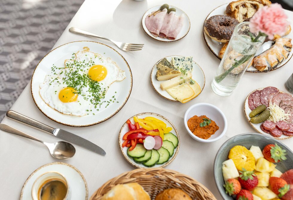Auf dem Frühstückstisch stehen Spiegeleier, Aufschnitt, Käse, Obst und Brot in einer hellen Restaurantatmosphäre.