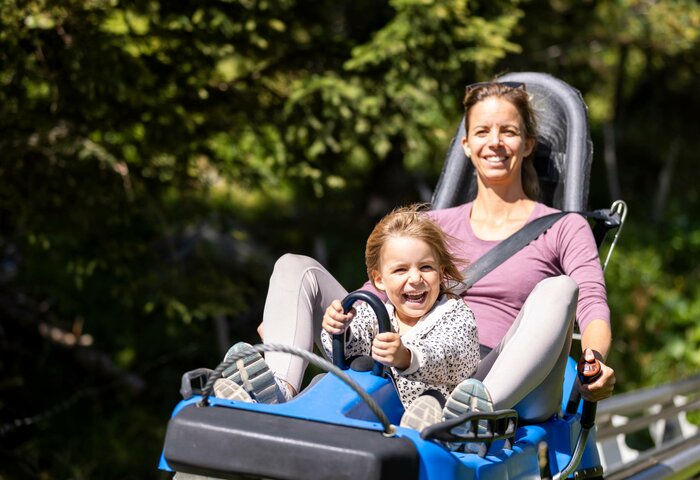 Eine Frau und ein Kind fahren gemeinsam auf einer Sommerrodelbahn und lächeln in der Sonne.