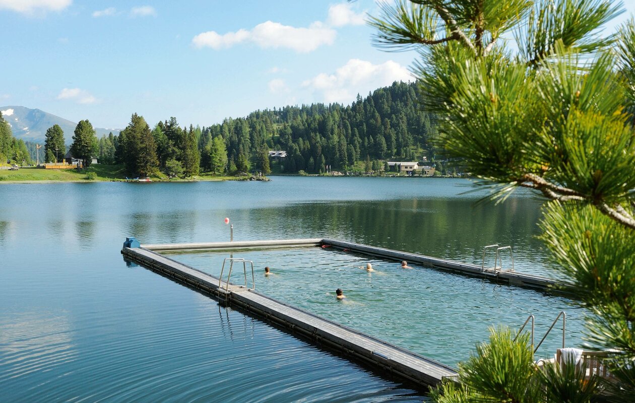 A heated lake pool with swimmers set in the lake, framed by forested mountains.