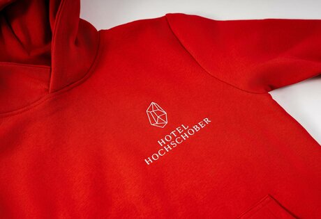 A red hoodie with the Hotel Hochschober logo printed on the chest is shown in close-up.