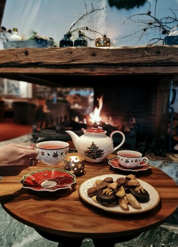 A tea service with pastries sits on a wooden table in front of a lit fireplace.
