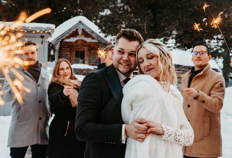 A bride and groom pose in the snow with sparklers while guests stand behind them.