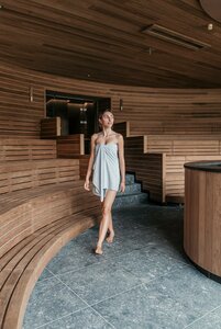 Bilder vom Wellnessurlaub mit Abwechslung in Österreich. Eine Frau in einem grauen Handtuch geht durch einen modernen Wellnessbereich mit Holzverkleidung und sanften, runden Formen. Die Umgebung strahlt Ruhe und Entspannung aus, mit Sitzgelegenheiten aus Holz und einem eleganten, minimalistischen Design.