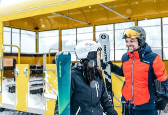 A skier and a skiier stand with their skis at a snowy mountain station.